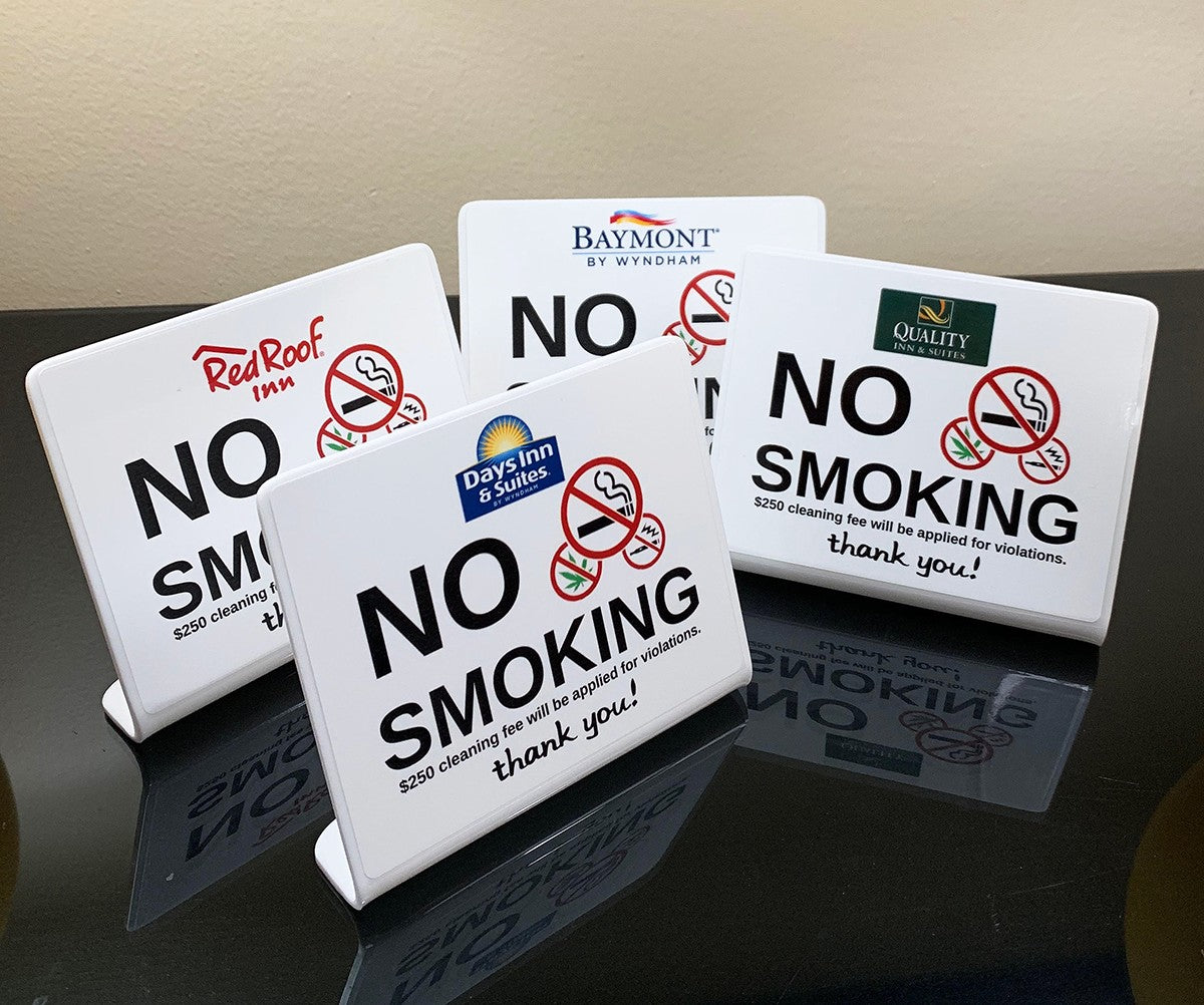 Four white plastic 'No Smoking' signs with red and black printed graphics and text, displayed on a glossy black reflective surface against a beige wall. The signs feature different hotel and resort logos including Red Roof Inn, Daysim Inn Suites, and Baymont by Wyndham.