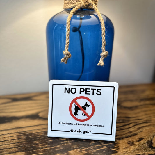A small white rectangular sign with a no-pets symbol and the text 'NO PETS thank you!' stands on a wooden surface in front of a blue glass bottle tied with a rope.