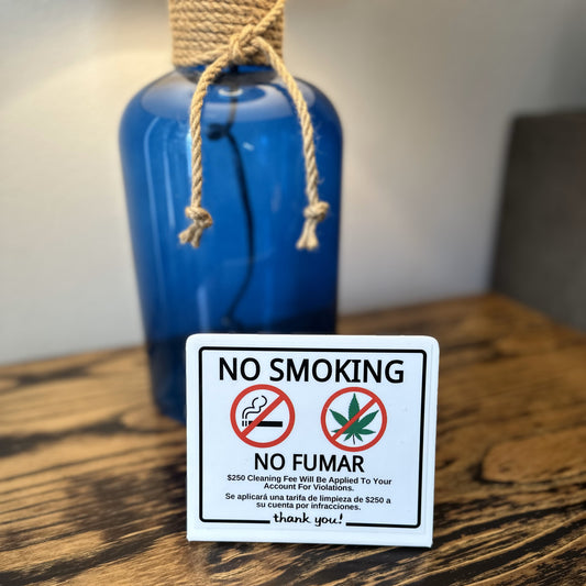 A square white 'NO SMOKING / NO FUMAR' sign with red prohibition symbols and black text, placed on a wooden table in front of a blue bottle with a rope neck wrap.