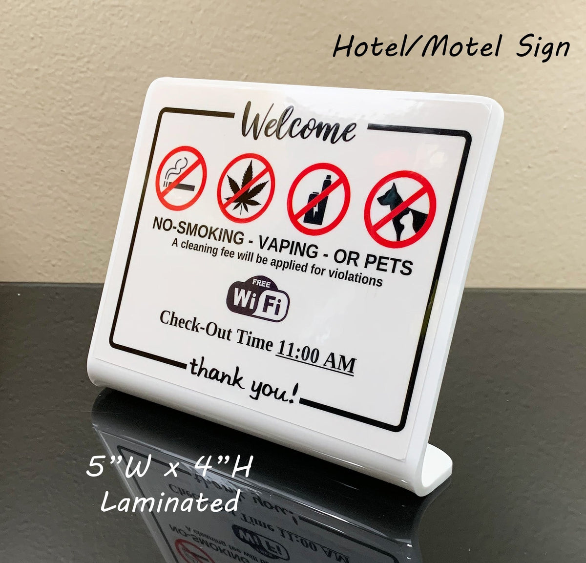 A white laminated hotel or motel welcome sign on a small stand, with black text and red prohibition icons for smoking, vaping, alcohol, and pets. The sign is photographed on a dark reflective surface against a beige wall, with overlaid text noting 'Hotel/Motel Sign' and dimensions '5" W x 4" H Laminated'.