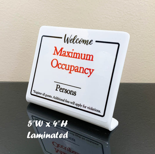 A white laminated acrylic maximum occupancy sign with red and black text, standing on a black reflective surface against a beige wall. White text overlay at the bottom left reads '5"W x 4"H Laminated'.