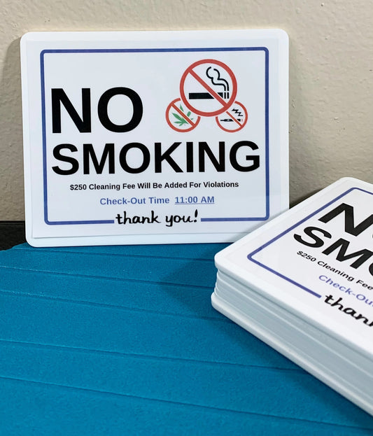 A square white 'NO SMOKING' sign with blue border lines, red prohibition symbols, and small black text, displayed upright on a blue surface with a stack of similar signs in the foreground.